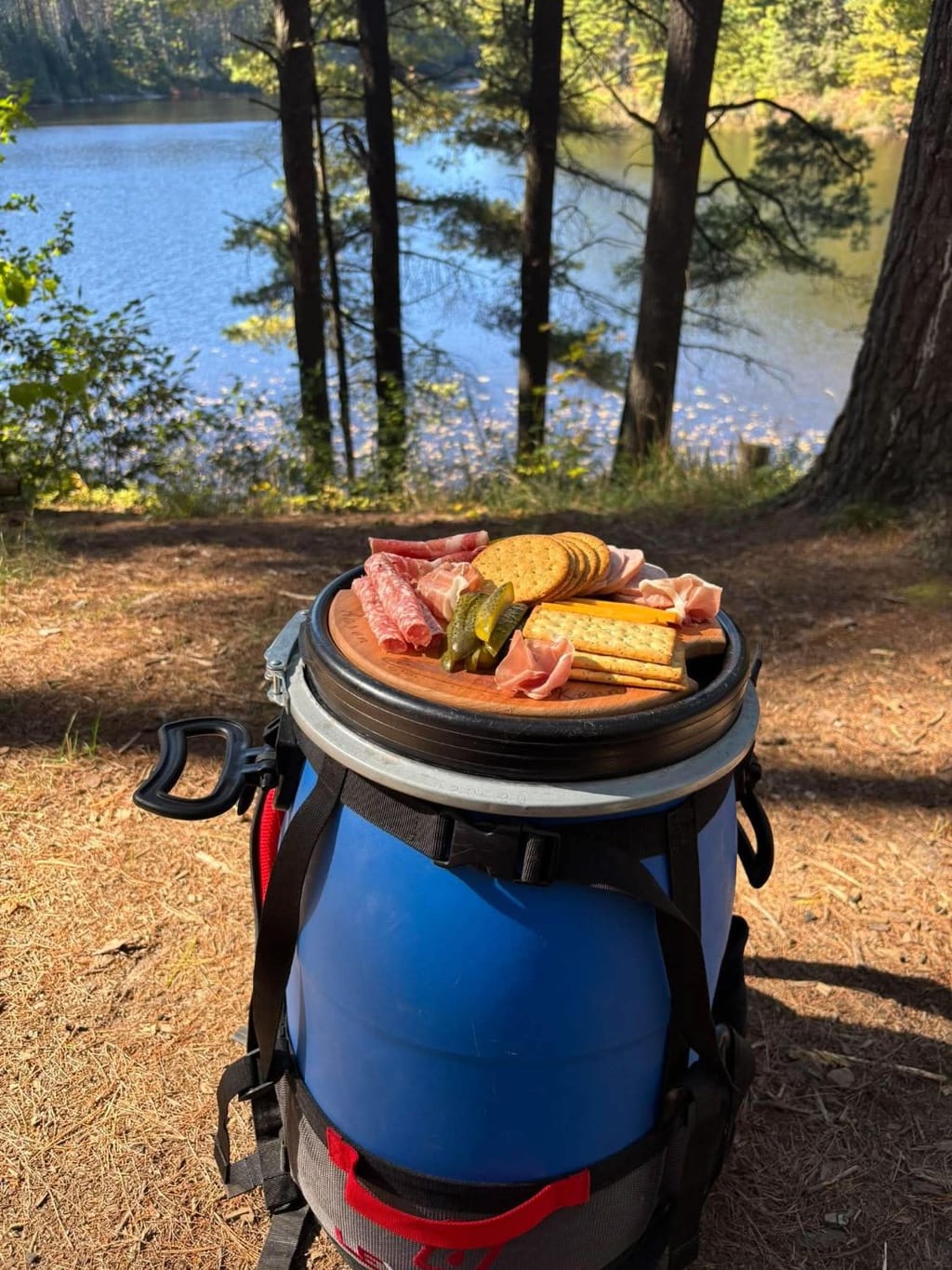 Barrel Buddy cutting board in a blue barrel lid with cured meats, crackers, cheese and pickles, overlooking a lake