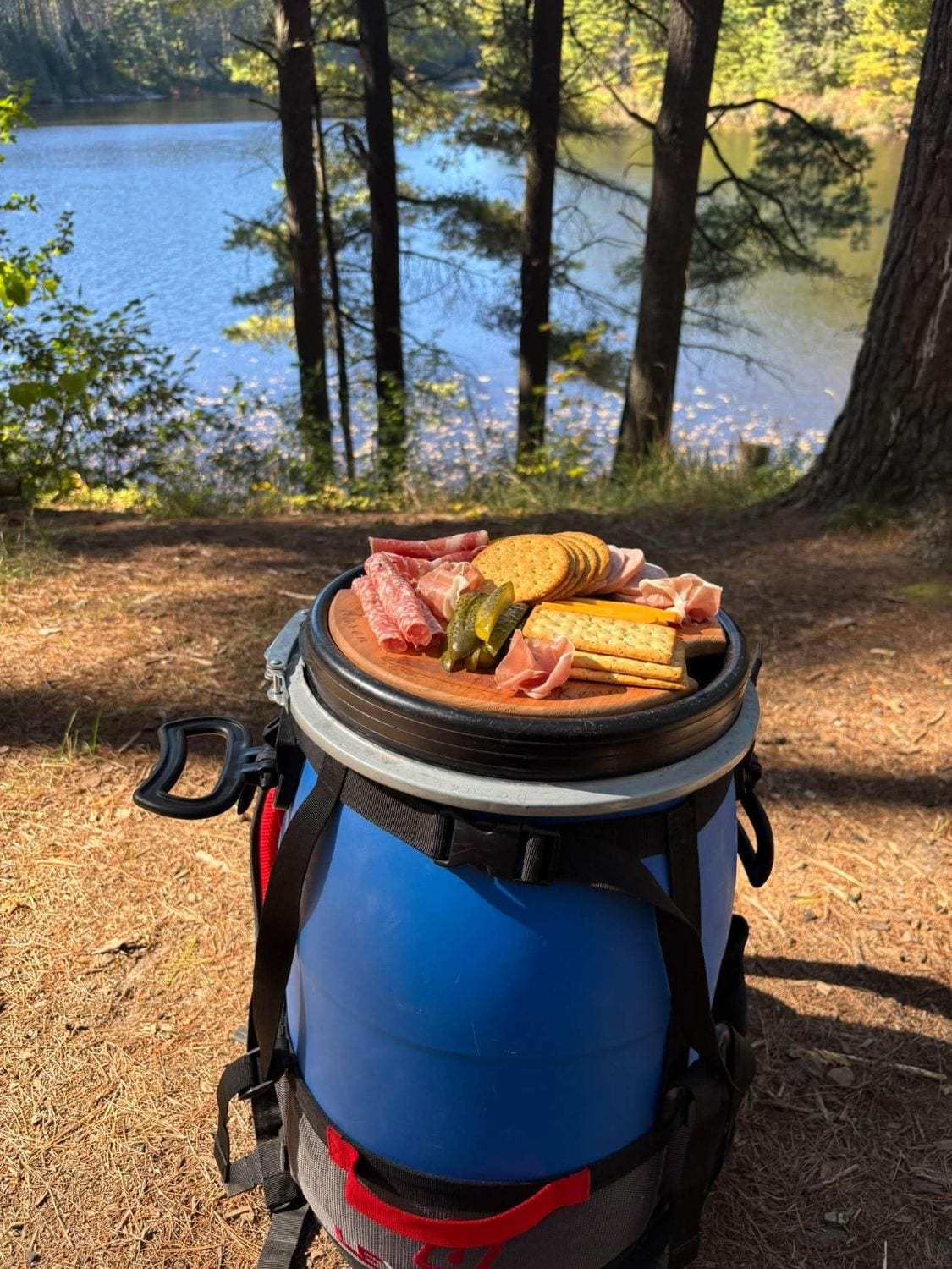 Barrel Buddy cutting board in a blue barrel lid with cured meats, crackers, cheese and pickles, overlooking a lake
